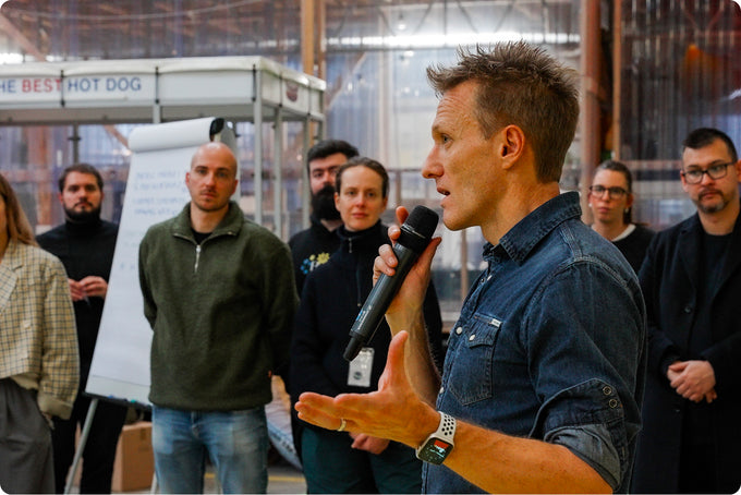 Man speaking into a microphone with a group of people listening in an indoor setting.