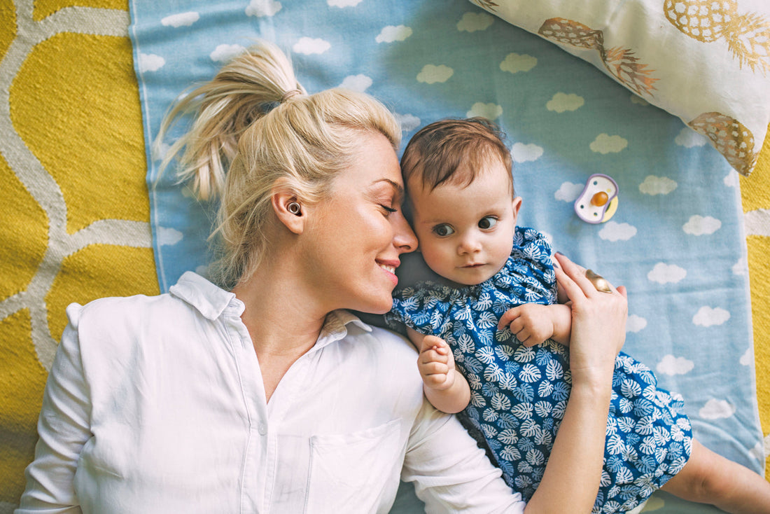 Image of a happy woman wearing Loop earplugs to manage her noise sensitivity and a baby laying on a blue and yellow coloured blanket