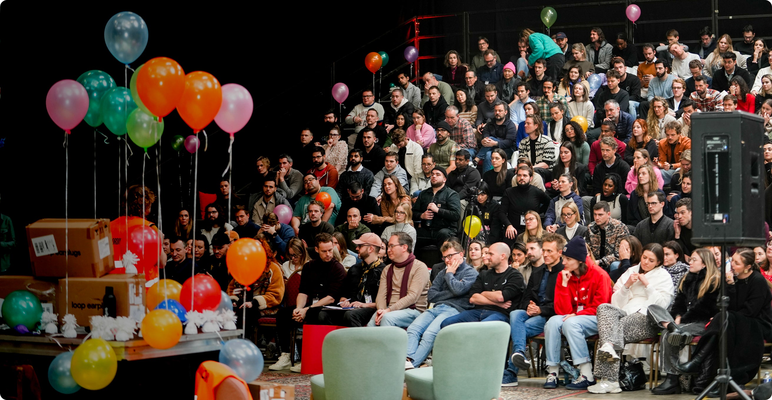 Audience in a theater with colorful balloons and a speaker.